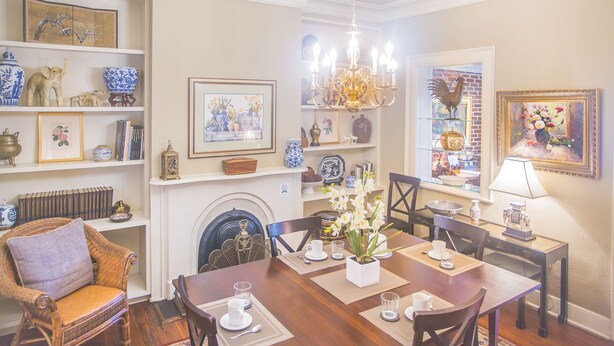 A dining area with a wooden table set for four, surrounded by chairs. There is a fireplace with a mantel, and shelves above holding various decorative items and framed artwork.