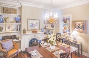 Un comedor con una mesa de madera preparada para cuatro personas, rodeada de sillas. Hay una chimenea con repisa, y estantes que exhiben diversos objetos decorativos y obras de arte enmarcadas.