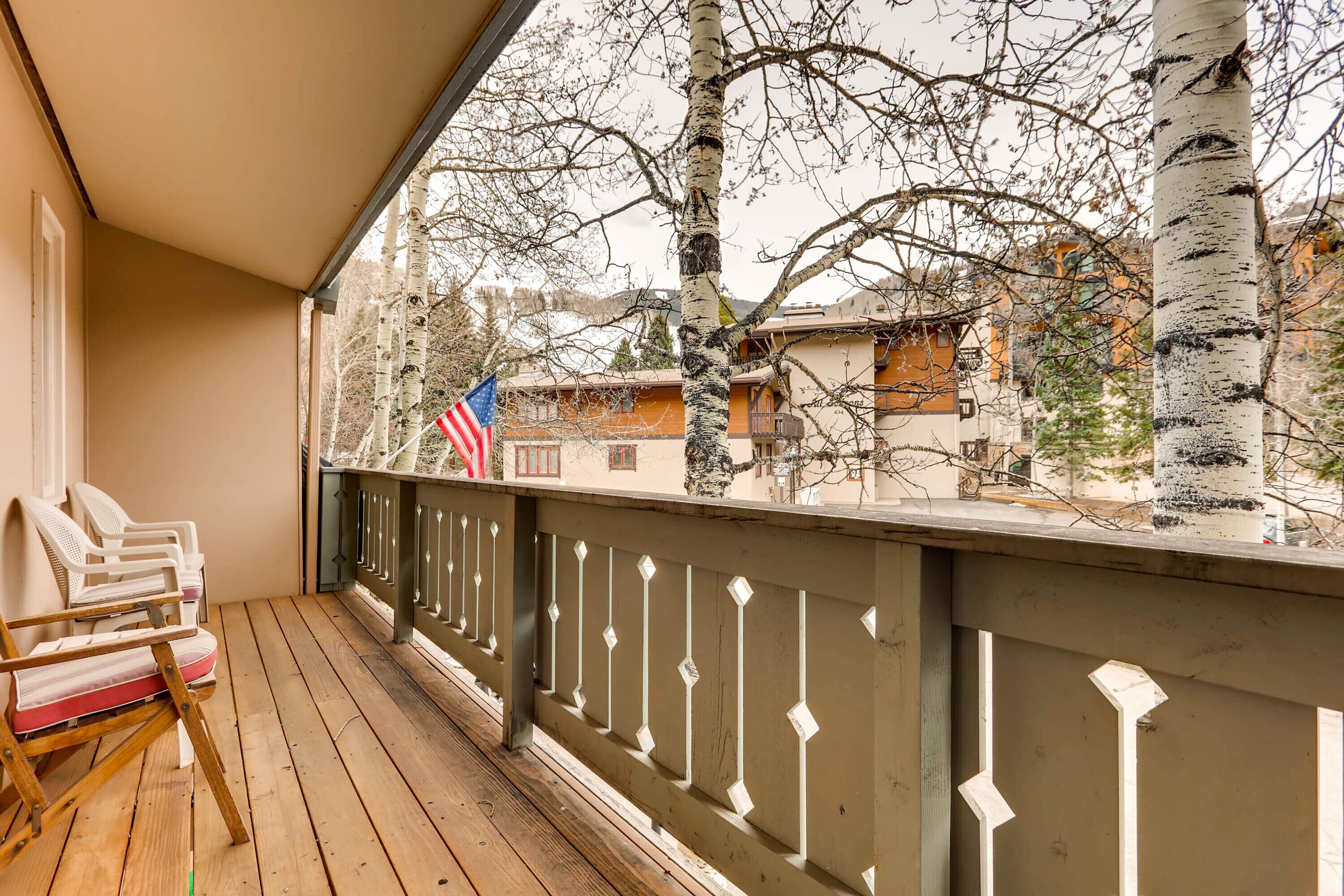 Une terrasse en bois avec des chaises, une rambarde et une vue sur les bâtiments et les arbres.