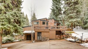 A modern two-story house with a balcony, surrounded by tall evergreen trees and a snow-covered ground.