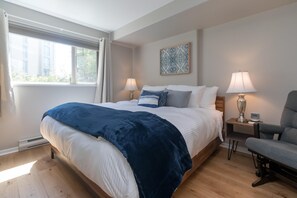 Ein Schlafzimmer mit einem Bett, zwei Nachttischen, einem Sessel, einem Fenster mit Vorhängen und einem Wandbild.