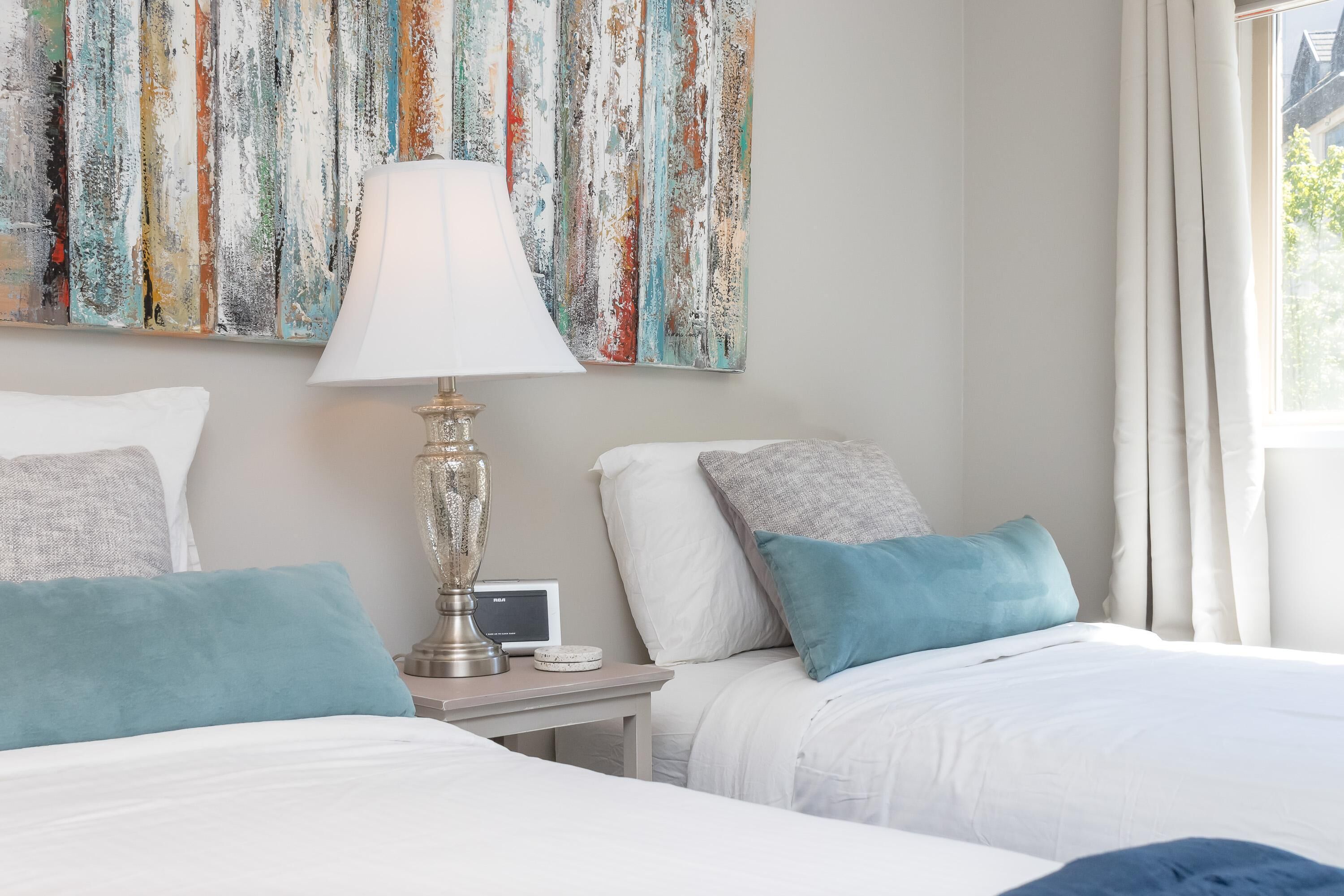 Une chambre d’hôtel avec deux lits, une table de chevet avec une lampe, une horloge et un tableau abstrait aux couleurs vives accroché au mur.