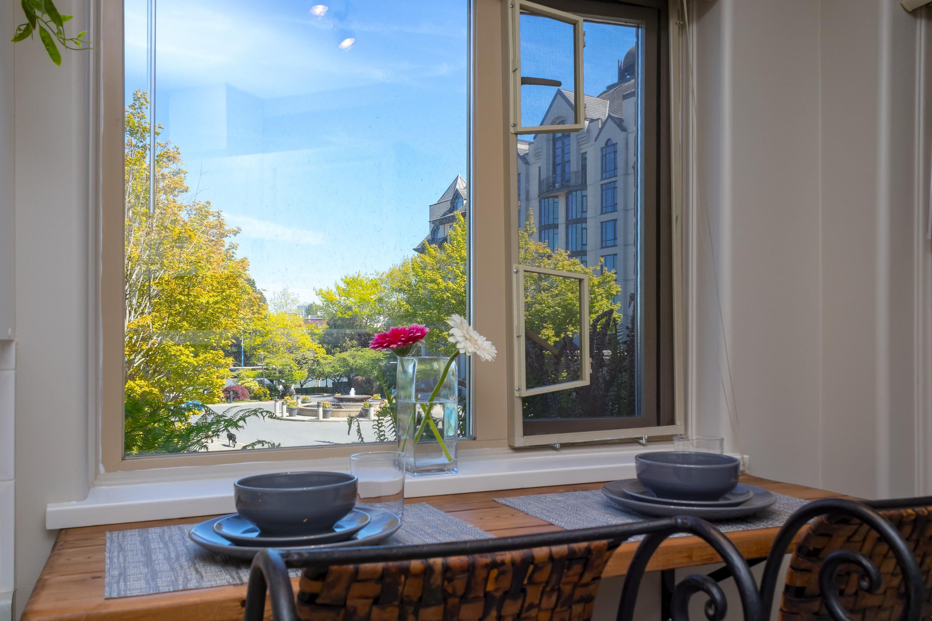 A dining area with a table set for two, a vase with flowers, and a view of a park and buildings through a window.