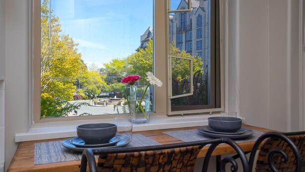 Spisestue med et bord dækket til to personer, en vase med blomster og udsigt til en park og bygninger gennem et vindue.
