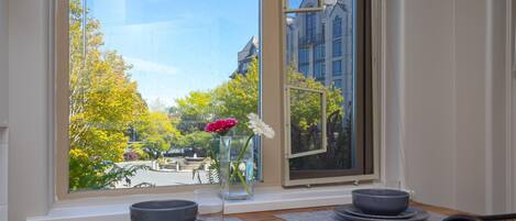 A dining area with a table set for two, a vase with flowers, and a view of a park and buildings through a window.