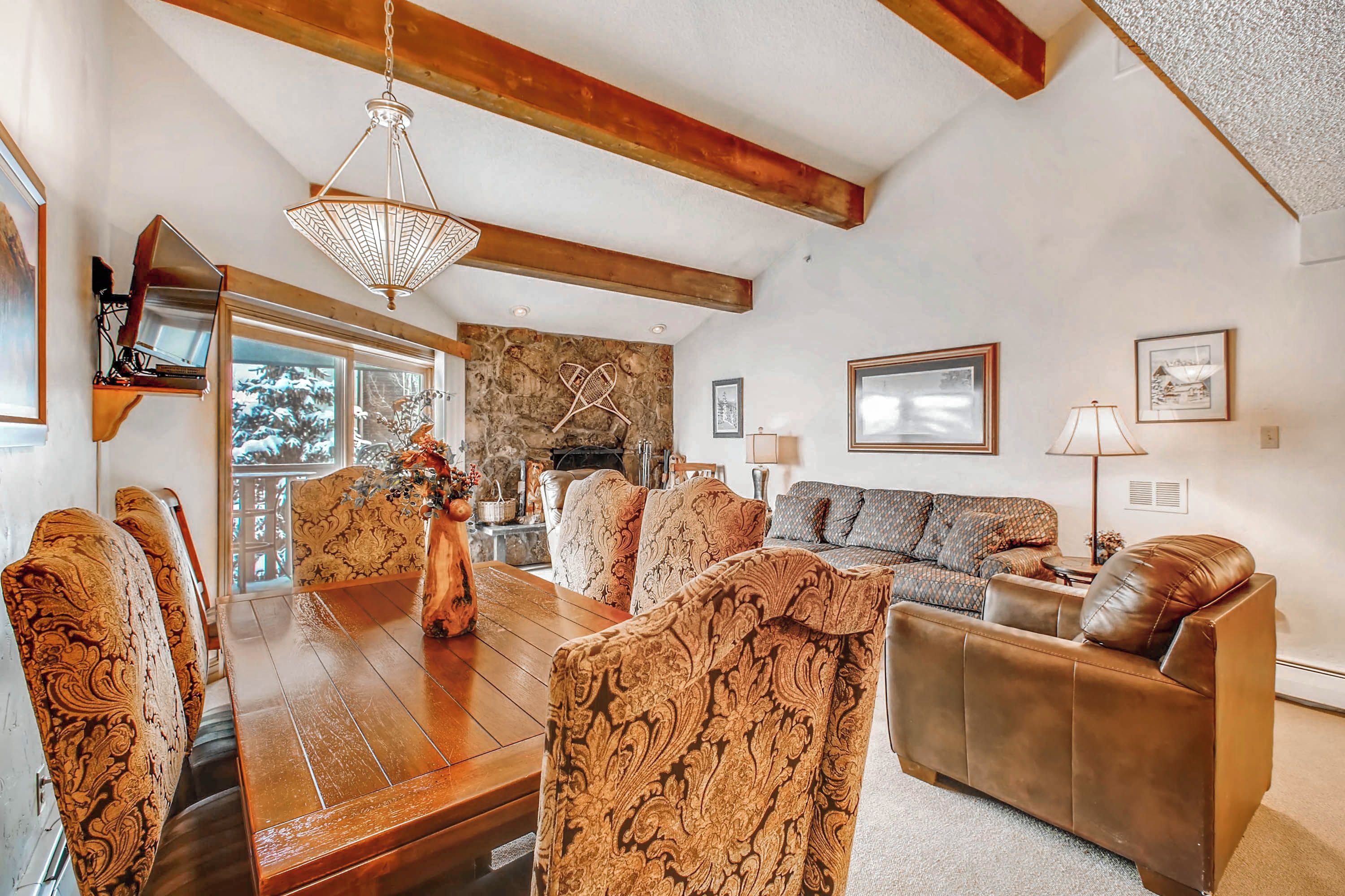 A spacious living room with a wooden dining table, patterned chairs, a brown leather sofa, and a stone fireplace.