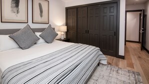 A bedroom with a large bed, grey pillows, a striped bedspread, a nightstand with a lamp, and a dark wooden wardrobe.