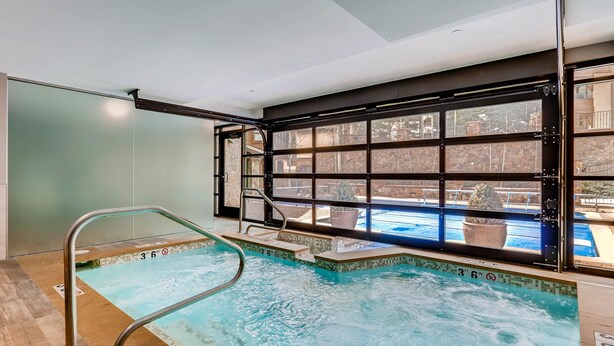 Ein Hallenbad mit klarem Wasser, einer Metallleiter und Blick auf ein Außenbecken durch große Fenster.