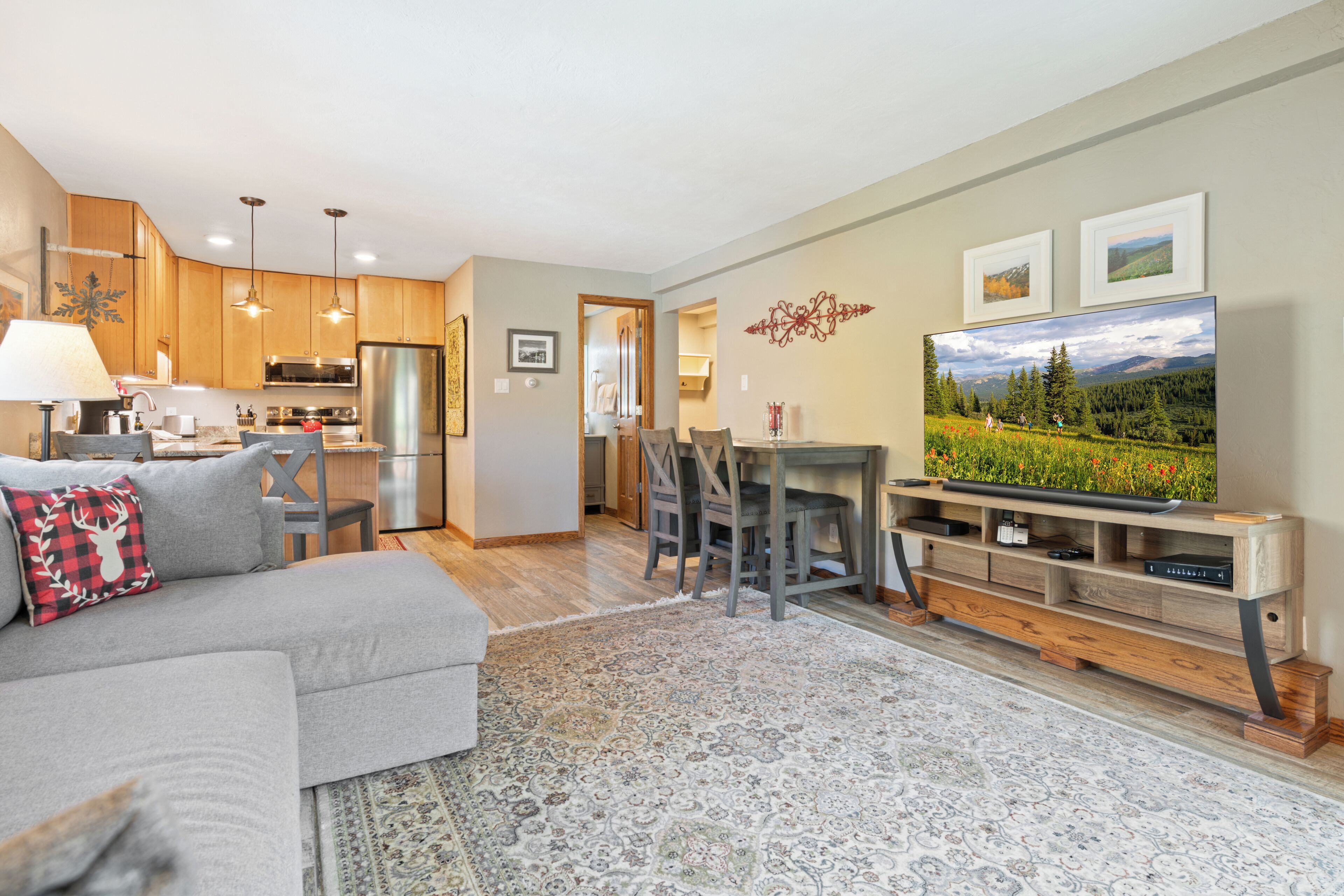 A modern living room with a grey sofa, a dining area with a wooden table and chairs, a kitchen with wooden cabinets and stainless steel appliances, and a large TV on the wall.