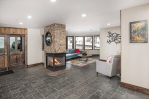 A modern living room with a stone fireplace, a grey sofa, and a large window offering a view of the outdoors. - Luxury Condo, Pool, Hot Tub, near Gondola | W108 (Vail)