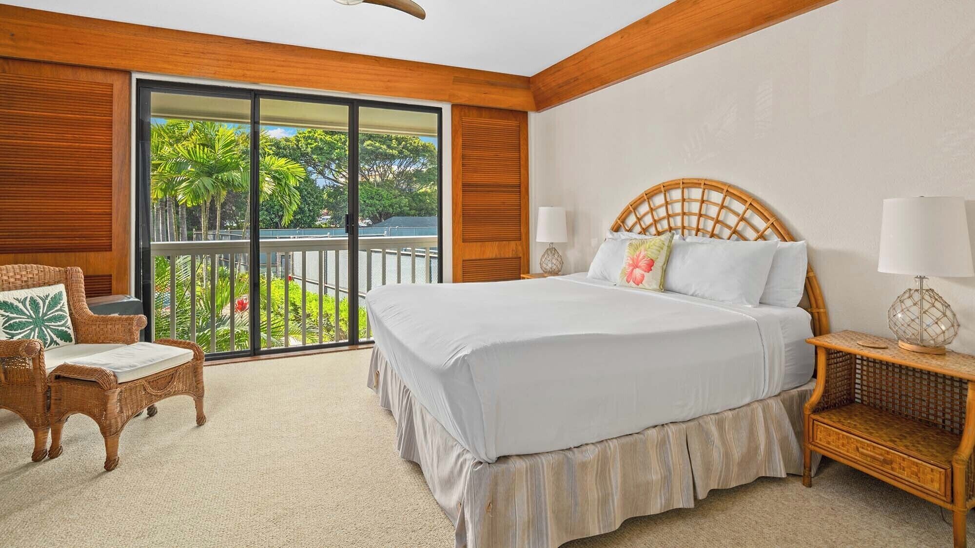 A bedroom with a large bed, a wicker chair, a nightstand with a lamp, and a sliding glass door leading to a balcony with greenery.
