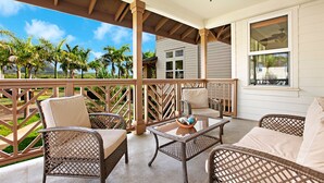 Un patio techado al aire libre con mobiliario de mimbre, una pequeña mesa y vistas a un jardín ajardinado y una casa al fondo.