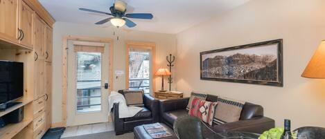 A cozy living room with a television, a glass coffee table, and a sofa. There is a ceiling fan, a door with a window, and a framed picture on the wall.