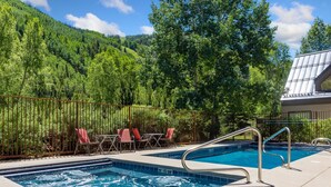 A swimming pool with metal railings, surrounded by a fence and red chairs, with a forested hillside in the background.