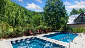 Una piscina con una escalera metálica, rodeada por una valla, con sillas y una mesa, y montañas al fondo.