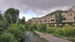 Una hilera de edificios de varios pisos con balcones, un río en primer plano y un paisaje montañoso al fondo.