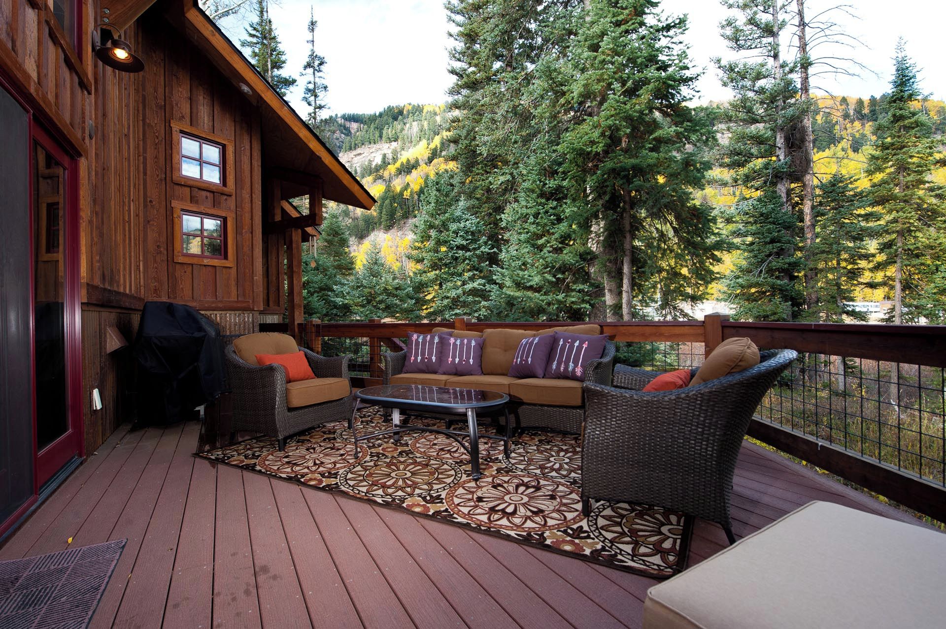 A wooden deck with outdoor furniture, a view of a forested mountain, and a cabin with a red door.