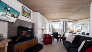 Ein Wohnzimmer mit Kamin, einem an der Wand befestigten Fernseher, einem roten Sessel, einem schwarzen Sofa und Blick auf die Berge durch große Fenster.