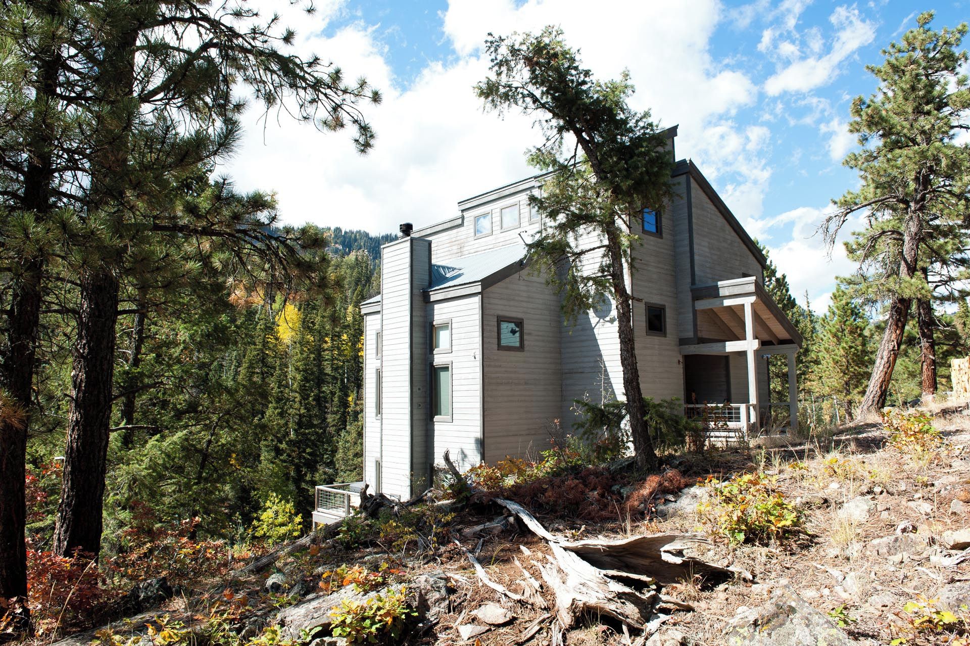 A two-story cabin with a covered porch and a small deck, surrounded by dense forest.
