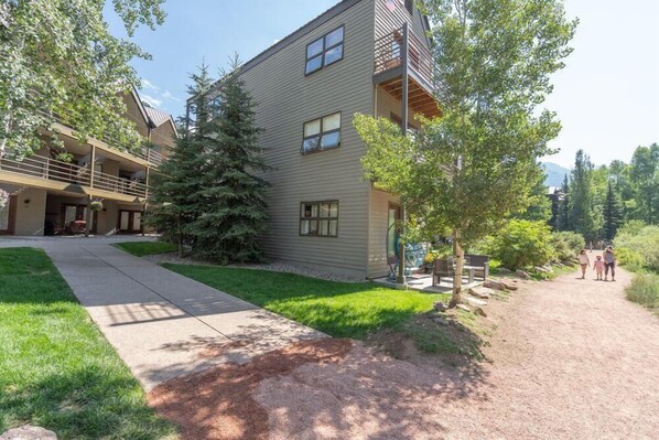 A two-story building with a balcony, a large tree in front, and a dirt path leading to a group of people.