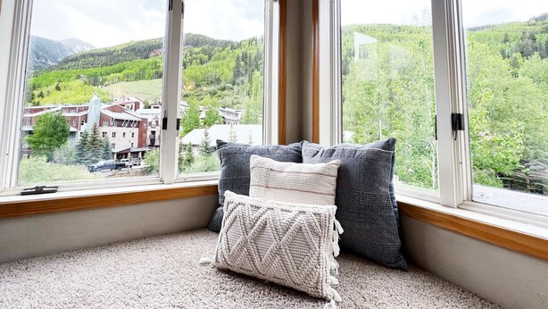 A room with a large window overlooking a green landscape and a small town. There are two pillows on the windowsill.
