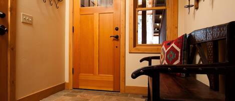 A wooden door with a glass panel, a stone-tiled floor, a bench with a patterned cushion, and a wall-mounted metal rack.