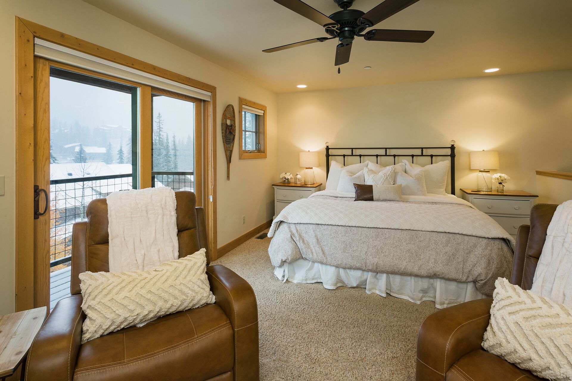A bedroom with a large bed, two nightstands, a ceiling fan, and a view of a snowy landscape through a sliding door.
