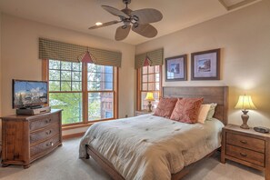 A bedroom with a bed, dresser, nightstands, lamps, ceiling fan, windows, and framed pictures on the wall.