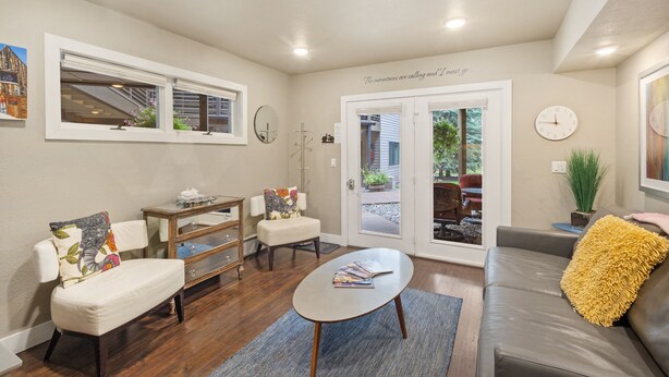 A living room with a sofa, armchair, coffee table, and a small side table. There is a door leading to an outdoor area with a view of a house and a clock on the wall. - Cozy Viking Lodge 105: One-bedroom escape with hot tub access (Telluride)