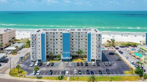 A multi-story hotel with a central blue tower, parking lot, and a beach view.