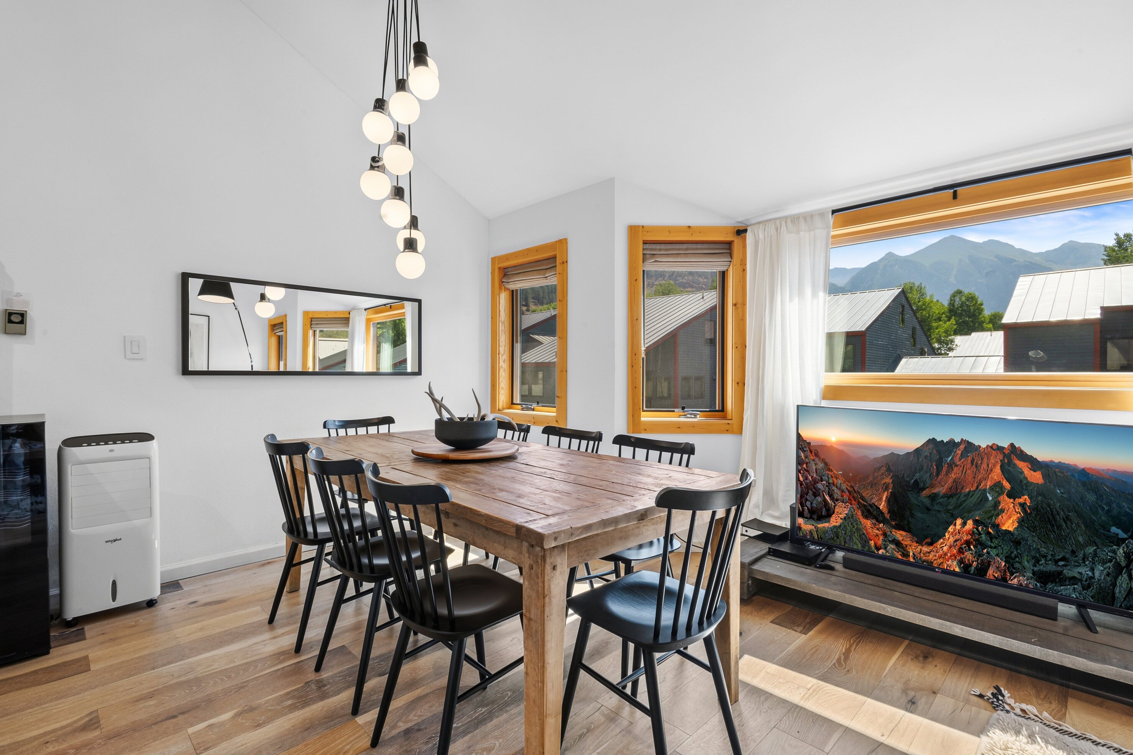 Un coin repas avec une table et des chaises en bois, une grande fenêtre donnant sur les montagnes et une cuisine moderne équipée d’un réfrigérateur et d’un four à micro-ondes.
