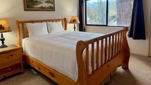Un dormitorio con una cama de madera, dos mesitas de noche, una ventana con vistas a árboles y un cuadro en la pared.