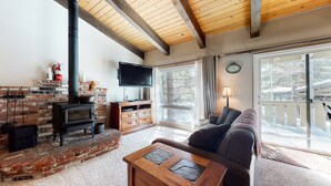 A cozy living room with a brick fireplace, a flat-screen TV, a comfortable sofa, and a large window offering a view of the outdoors. - Cozy Mountain Home, Shuttle Route, Walk to Bowling Alley & Washer/Dryer (Mammoth Lakes)
