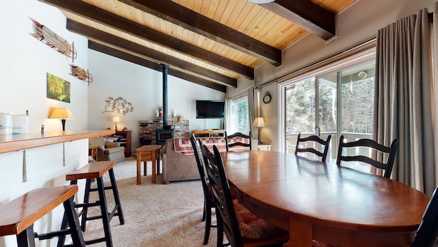 A cozy living room with a fireplace, wooden ceiling beams, a dining table with chairs, and a large window with curtains. - Cozy Mountain Home, Shuttle Route, Walk to Bowling Alley & Washer/Dryer (Mammoth Lakes)