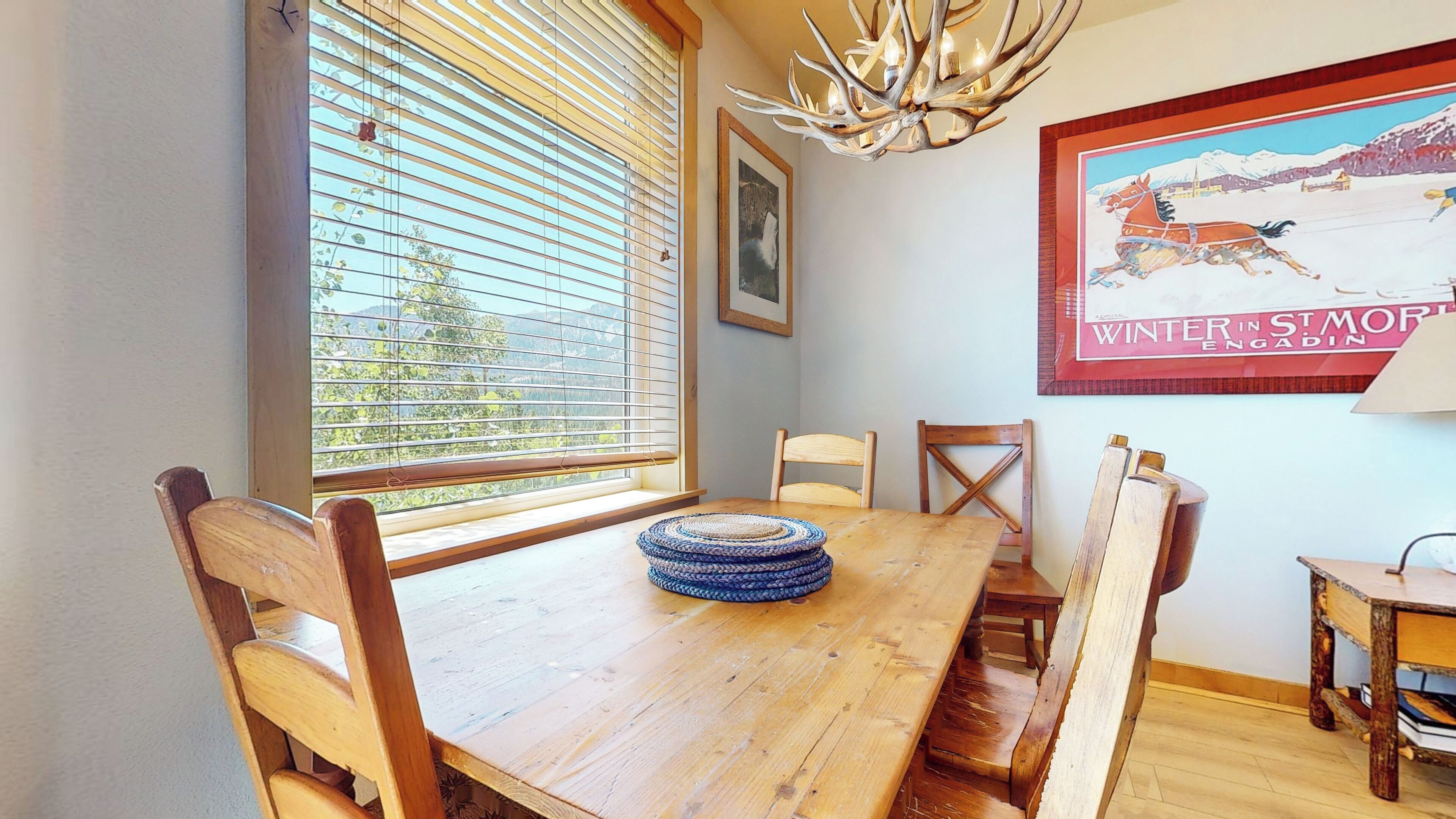 A wooden dining table with chairs, a woven placemat, a framed picture, and a chandelier.