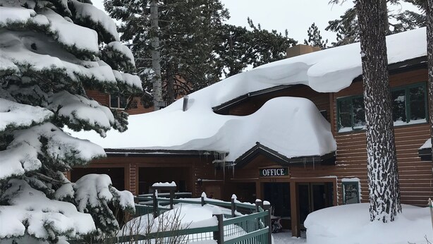 Uma cabana de madeira com uma placa indicando 'OFFICE' na entrada, rodeada por árvores cobertas de neve e uma área cercada.