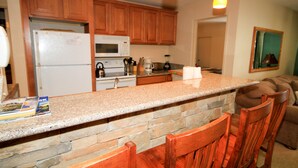 A kitchen with wooden cabinets, a granite countertop, a refrigerator, a microwave, and a coffee maker. There are three wooden chairs at the counter.