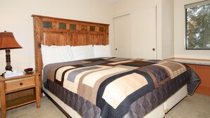 A bedroom with a wooden headboard, a quilted bedspread, a nightstand with a lamp, and a window with a view of trees.