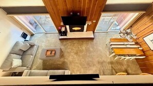 A modern living room with a wooden ceiling, a flat-screen TV, a grey sofa, a coffee table, and a dining area with a table and chairs. - Mammoth Ski & Racquet Club #120 - Pet Friendly (Mammoth Lakes)