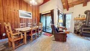 Una habitación acogedora con paredes de madera, una mesa de comedor con sillas, chimenea y un ventanal con cortinas.