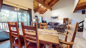 Una mesa de comedor de madera con sillas, un sofá y una televisión en una sala de estar con chimenea de piedra y un ventanal grande.