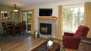 Una sala de estar con chimenea de piedra, un televisor montado encima, un comedor con mesa y sillas, y un sillón rojo.