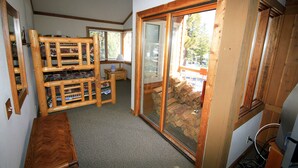 A room with a wooden bench, a small table, and a large sliding glass door leading to an outdoor area with a view of trees. - Mountainback #63, Loft, Den (Mammoth Lakes)