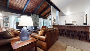 A cozy living room with a brown leather sofa, a wooden coffee table, and a stone wall. A kitchen with white cabinets and a bar area is visible in the background. - Mountain Rustic, Pool & Tennis, Near Dining, Rec Center, Bowling & Trails (Mammoth Lakes)