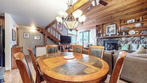 Un interior acogedor de cabaña con una mesa de comedor de madera, sillas, una chimenea y una escalera que lleva a un altillo.