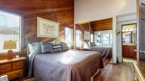 Un dormitorio con una cama grande, piso de madera, cómoda, mesita de noche, lámpara y ventana con persianas.