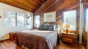 A cozy bedroom with a wooden ceiling, a large bed with patterned bedding, a bedside table with a lamp, and framed artwork on the wall.