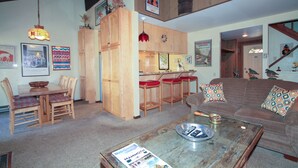 Ein gemütliches Wohnzimmer mit einem Holzesstisch, rot gepolsterten Stühlen, einem braunen Sofa mit gemusterten Kissen und einem Couchtisch mit einer Zeitschrift.