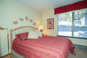 A bedroom with a wooden bed, a red and white patterned bedspread, a bedside lamp, and a window with blinds.