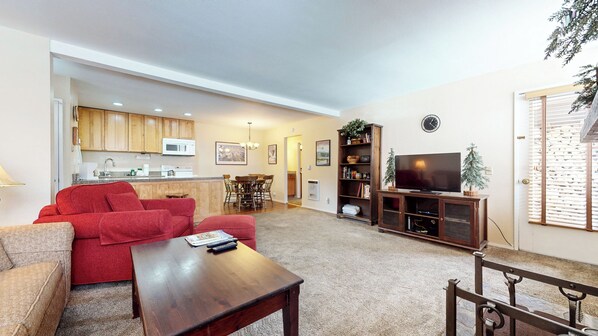 A living room with a red sofa, a wooden coffee table, a TV on a wooden stand, and a kitchen in the background. - Homey & Updated Condo near The Village, Private Balcony. Hot Tub On-site (Mammoth Lakes)
